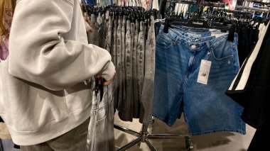 Moda bir giyim mağazasında kot pantolon almak için kapüşon giyen genç bir kadın. Moda alışverişi, modern yaşam tarzı, gündelik moda, ergen modası, perakende deneyimi. Bir Giyim Mağazasında Çok Giysi Askısı Tutan Kız, Alışveriş Konsepti, Fa