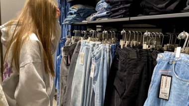 Moda bir giyim mağazasında kot pantolon almak için kapüşon giyen genç bir kadın. Moda alışverişi, modern yaşam tarzı, gündelik moda, ergen modası, perakende deneyimi. Kıyafet Mağazasında Çoklu Elbise Askısı Tutan Kız, Alışveriş Konsepti