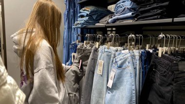 Moda bir giyim mağazasında kot pantolon almak için kapüşon giyen genç bir kadın. Moda alışverişi, modern yaşam tarzı, gündelik moda, ergen modası, perakende deneyimi. Kıyafet Mağazasında Çoklu Elbise Askısı Tutan Kız, Alışveriş Konsepti