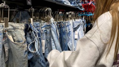 Moda bir giyim mağazasında kot pantolon almak için kapüşon giyen genç bir kadın. Moda alışverişi, modern yaşam tarzı, gündelik moda, ergen modası, perakende deneyimi. Kıyafet Mağazasında Çoklu Elbise Askısı Tutan Kız, Alışveriş Konsepti