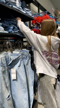Moda bir giyim mağazasında kot pantolon almak için kapüşon giyen genç bir kadın. Moda alışverişi, modern yaşam tarzı, gündelik moda, ergen modası, perakende deneyimi. Kıyafet Mağazasında Çoklu Elbise Askısı Tutan Kız, Alışveriş Konsepti