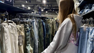 Moda bir giyim mağazasında kot pantolon almak için kapüşon giyen genç bir kadın. Moda alışverişi, modern yaşam tarzı, gündelik moda, ergen modası, perakende deneyimi. Kıyafet Mağazasında Çoklu Elbise Askısı Tutan Kız, Alışveriş Konsepti