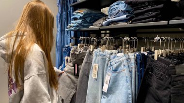 Moda bir giyim mağazasında kot pantolon almak için kapüşon giyen genç bir kadın. Moda alışverişi, modern yaşam tarzı, gündelik moda, ergen modası, perakende deneyimi. Kıyafet Mağazasında Çoklu Elbise Askısı Tutan Kız, Alışveriş Konsepti