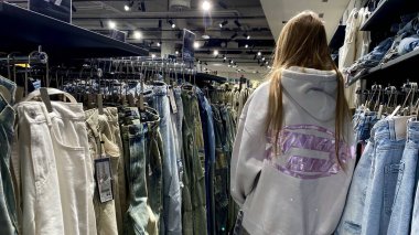 Moda bir giyim mağazasında kot pantolon almak için kapüşon giyen genç bir kadın. Moda alışverişi, modern yaşam tarzı, gündelik moda, ergen modası, perakende deneyimi. Kıyafet Mağazasında Çoklu Elbise Askısı Tutan Kız, Alışveriş Konsepti