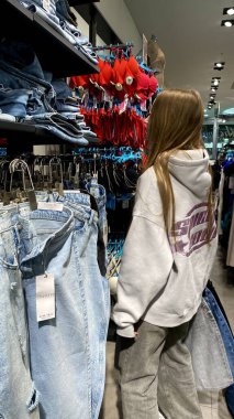 Moda bir giyim mağazasında kot pantolon almak için kapüşon giyen genç bir kadın. Moda alışverişi, modern yaşam tarzı, gündelik moda, ergen modası, perakende deneyimi. Kıyafet Mağazasında Çoklu Elbise Askısı Tutan Kız, Alışveriş Konsepti