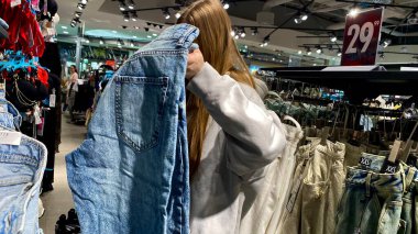 Moda bir giyim mağazasında kot pantolon almak için kapüşon giyen genç bir kadın. Moda alışverişi, modern yaşam tarzı, gündelik moda, ergen modası, perakende deneyimi. Kıyafet Mağazasında Çoklu Elbise Askısı Tutan Kız, Alışveriş Konsepti