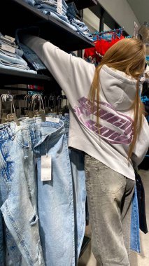 Moda bir giyim mağazasında kot pantolon almak için kapüşon giyen genç bir kadın. Moda alışverişi, modern yaşam tarzı, gündelik moda, ergen modası, perakende deneyimi. Kıyafet Mağazasında Çoklu Elbise Askısı Tutan Kız, Alışveriş Konsepti
