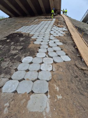 Köprü yenilenmesi ve modern bir inşaat sahasında kaldırım döşemesi sergilenen yüksek kaliteli bir fotoğraf. İşçiler aktif olarak yol çalışması yapıyor, şehir altyapısını geliştiriyorlar. Olay yerinde inşaat ekipmanları, malzemeler ve
