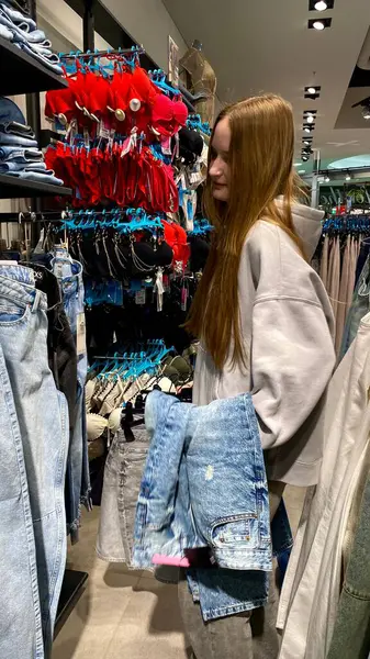 Moda bir giyim mağazasında kot pantolon almak için kapüşon giyen genç bir kadın. Moda alışverişi, modern yaşam tarzı, gündelik moda, ergen modası, perakende deneyimi. Kıyafet Mağazasında Çoklu Elbise Askısı Tutan Kız, Alışveriş Konsepti