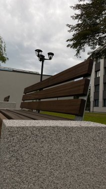 Modern bir konut binasının peyzajlı avlusunda yer alan beton desteklerden ve ahşap oturma yüzeyinden oluşan çağdaş bir tezgah. Bu bank konfor, rahatlama ve kentsel aes için tasarlanmış iyi korunmuş bir kamu alanının parçasıdır.