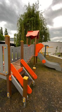 Renkli bir kaydırak, tırmanma merdiveni, küçük ahşap oyun evi ve köprü yapısı olan modern açık hava çocukları için oyun alanı. Kamu parkında ya da eğlence alanında geçen bu ekipman fiziksel aktiviteyi, hayal gücünü ve sosyal etkileşimi teşvik eder..