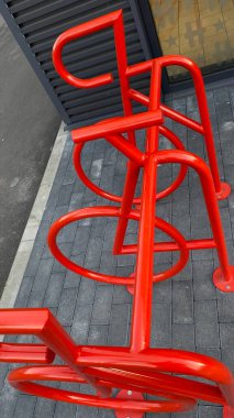 Bisiklet şeklinde tasarlanmış parlak kırmızı bir bisiklet askısı modern bir binanın yanındaki kaldırımda duruyor. Bu kentsel altyapı unsuru işlevselliği yaratıcı kamu tasarımı ile birleştirir. Görüntü temiz, organize bir şehir ortamını yakalıyor.