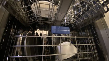 Bir kız modern bir bulaşık makinesine temiz ve çağdaş bir mutfakta tabak yüklüyor. İçi granit kaplama, beyaz dolaplar ve saksı bitkisi içeriyor. Görüntü günlük ev işlerini, mutfak düzenini ve kullanım şeklini gösteriyor.