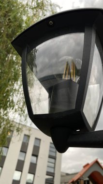 Metal direkli ve entegre ışıklandırmalı modern kentsel sokak lambası, yeşil yeşillik bir zemin ve çağdaş bir konut binasına karşı çekilmiş. Kompozisyon, kentsel altyapı, doğa ve