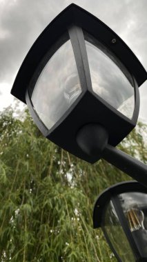 Metal direkli ve entegre ışıklandırmalı modern kentsel sokak lambası, yeşil yeşillik bir zemin ve çağdaş bir konut binasına karşı çekilmiş. Kompozisyon, kentsel altyapı, doğa ve