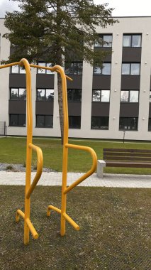 Modern bir konut avlusuna yerleştirilmiş sağlam metal paralel barlar, bir çocuk oyun parkının yakınında yer alıyor. Vücut ağırlık egzersizleri ve sokak fitness rutinleri için tasarlanan ekipman, fiziksel gelişimi destekliyor ve aktif bir kentsel yaşamı teşvik ediyor.