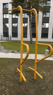 Modern bir konut avlusuna yerleştirilmiş sağlam metal paralel barlar, bir çocuk oyun parkının yakınında yer alıyor. Vücut ağırlık egzersizleri ve sokak fitness rutinleri için tasarlanan ekipman, fiziksel gelişimi destekliyor ve aktif bir kentsel yaşamı teşvik ediyor.