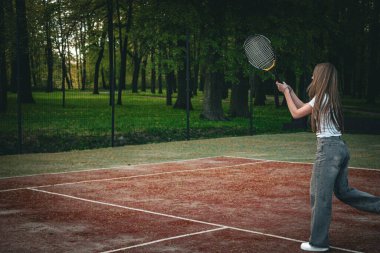 Genç bir kız açık hava kil kortunda tenis topuna raketle vuruyor. Saray yeşil bir orman ve tel örgü ile çevrili. Sıradan bir kot pantolon ve beyaz bir tişört giyip bahar etkinliğinin tadını çıkarıyor.. 