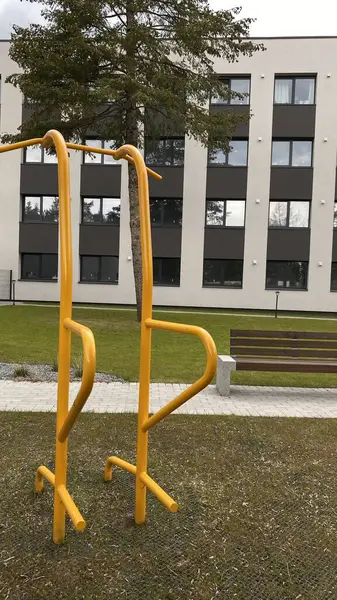 Modern bir konut avlusuna yerleştirilmiş sağlam metal paralel barlar, bir çocuk oyun parkının yakınında yer alıyor. Vücut ağırlık egzersizleri ve sokak fitness rutinleri için tasarlanan ekipman, fiziksel gelişimi destekliyor ve aktif bir kentsel yaşamı teşvik ediyor.