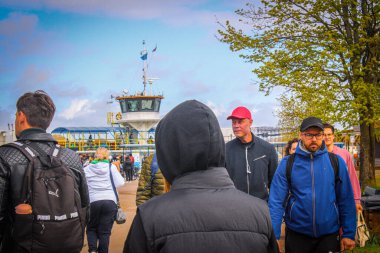 İnsanlar bir bahar günü feribot terminaline doğru yürüyorlar. Sıradan bir kalabalık feribota biniyor. Görünür gemi kontrol kulesi ve arka planda bayraklar var..