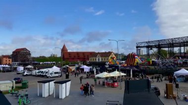 Litvanya 'daki Klaipeda Limanı' nda halka açık bir etkinlik. İnsanlar su kenarında bisiklet sürüyor ve aile dostu bir atmosferin tadını çıkarıyorlar. Rıhtım boyunca şişirilebilir çekimler beyaz çadırlar ve market tezgahları kuruluyor. Yatlar demirli.