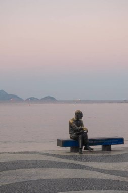 Rio de Janeiro, Brezilya, 06-07-2023: Brezilyalı şair Carlos Drummond de Andrade 'nin (1902-1987) heykelinin bulunduğu Copacabana plajında gün batımı ve onun tezgahtaki şiiri ve deniz üzerine yazılmış bir şehir