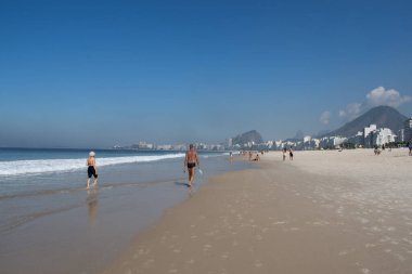 Rio de Janeiro, Brezilya, 06-07-2023: Copacabana plajının yanındaki Leme plajında yürüyen insanlar, şehrin gökdelenleri ve gökdelenleri Avenida Atlantica boyunca (Atlantik Bulvarı))