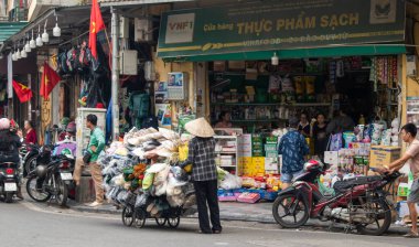 Hanoi, Vietnam, 11-06-2024: Geleneksel konik konik şapka takan Vietnamlı kadın Non La 'da araç gereç satıyor ve malları şehrin kalabalık caddelerinde taşıyarak markete gidiyor.