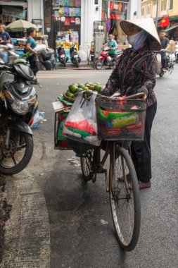 Hanoi, Vietnam, 11-06-2024: Geleneksel konik konik şapka takan ve meyve satan Vietnamlı bir kadın bisikletini şehrin kalabalık caddelerinde pazara doğru sürüyor.