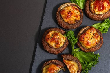 Pişmiş mantar kapları yenmeye hazır. Şampiyonlar, baharatlar, sebzeler, peynir. Hizmet edilen bölüm, modaya uygun sert ışık, karanlık gölge. Siyah taş arkaplan, üst görünüm