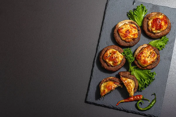 stock image Baked stuffed mushroom caps ready for eating. Champignons, spices, vegetables, cheese. Served portion, trendy hard light, dark shadow. Black stone concrete background, top view