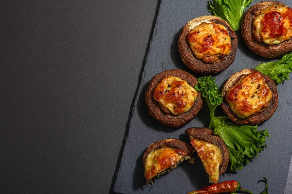 stock image Baked stuffed mushroom caps ready for eating. Champignons, spices, vegetables, cheese. Served portion, trendy hard light, dark shadow. Black stone concrete background, top view