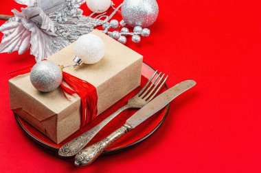 Noel sofrasında kırmızı ve gümüş renkli bir hediye kutusu var. Yeni yıl arkaplanı, seramik tabak ve geleneksel dekor. Düz konum, el yapımı tasarım, kopyalama alanı