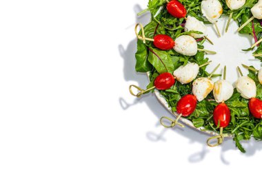 Şarküteri çelengi beyaz arka planda izole edilmiş. Mozzarella, vişneli domates ve şiş roka. Modaya uygun atıştırmalıklar, şenlikli vejetaryen yemekleri, modaya uygun sert ışık, karanlık gölge, üst manzara.