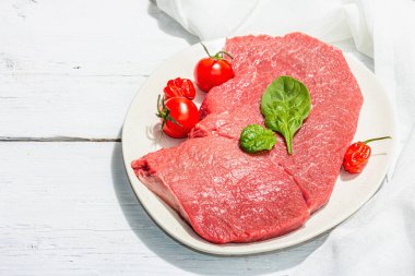 Raw veal steak with spices and cherry tomatoes. A trendy hard light, dark shadow, white wooden background, flat lay, copy space
