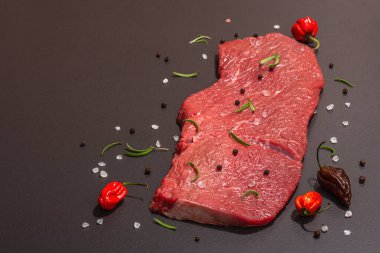 Raw veal steak with spices, rosemary and chili pepper. A trendy hard light, dark shadow, black stone concrete background, flat lay, copy space