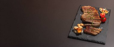 Well done roasting veal steak with sauce, rosemary and grilled garlic. A trendy hard light, dark shadow, black stone concrete background, flat lay, banner