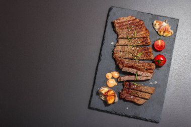 Well done roasting veal steak with sauce, rosemary and grilled garlic. A trendy hard light, dark shadow, black stone concrete background, flat lay, top view