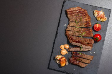 Well done roasting veal steak with sauce, rosemary and grilled garlic. A trendy hard light, dark shadow, black stone concrete background, flat lay, top view