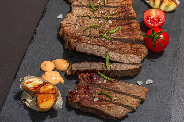Well done roasting veal steak with sauce, rosemary and grilled garlic. A trendy hard light, dark shadow, black stone concrete background, flat lay, close up