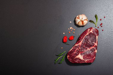 Raw Ribeye steak with spices and herbs. Dry aged meat, traditional food for grill. A trendy hard light, dark shadow, black stone concrete background, flat lay, top view