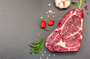 Raw Ribeye steak with spices and herbs. Dry aged meat, traditional food for grill. Black stone concrete background, flat lay, top view