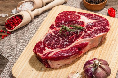 Raw Ribeye steak with spices and herbs. Dry aged meat, traditional food for grill. A trendy hard light, dark shadow, old wooden background, flat lay, close up