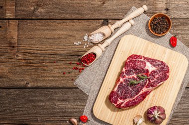 Raw Ribeye steak with spices and herbs. Dry aged meat, traditional food for grill. A trendy hard light, dark shadow, old wooden background, flat lay, top view