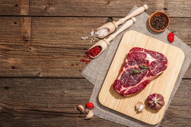 Raw Ribeye steak with spices and herbs. Dry aged meat, traditional food for grill. A trendy hard light, dark shadow, old wooden background, flat lay, top view