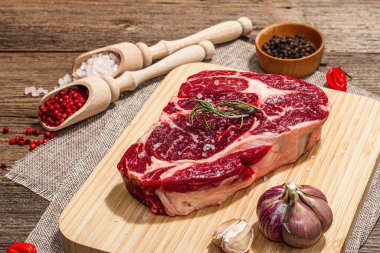 Raw Ribeye steak with spices and herbs. Dry aged meat, traditional food for grill. A trendy hard light, dark shadow, old wooden background, flat lay, close up