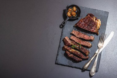 Well done roasting Ribeye steak with caramelized garlic and vintage cutlery. Trendy hard light, dark shadow, black stone concrete background, top view