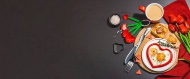 Heart-shaped fried egg served with toasted bread and spices on a plate. Romantic art food idea for Valentine's breakfast. Hard light, dark shadow, black stone concrete background, banner format