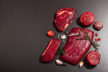 Set of various steaks with classic spices and herbs. Fresh raw meat cuts includes ribeye, eye round, flank and striploin steaks. Black stone concrete background, flat lay, top view
