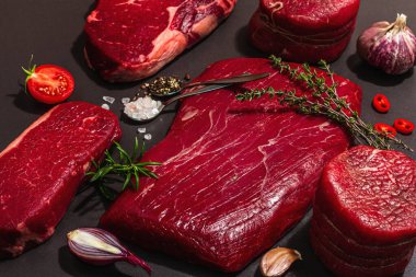 Set of various steaks with classic spices and herbs. Fresh raw meat cuts includes ribeye, eye round, flank and striploin steaks. Black stone concrete background, flat lay, close up